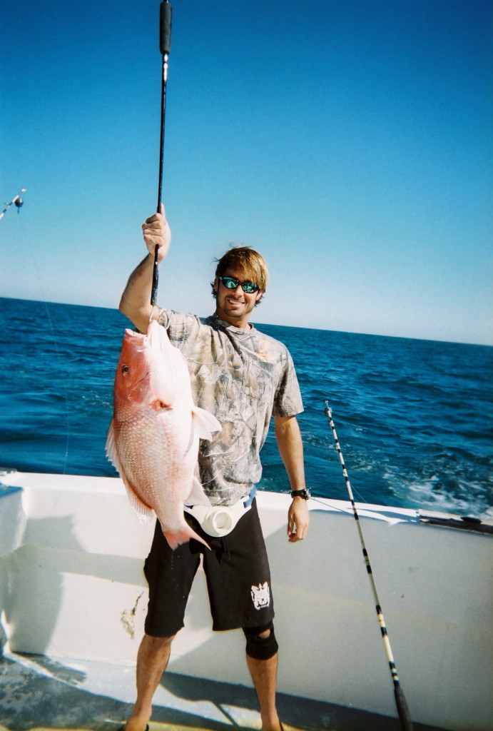 A smiling angler in shorts and sunglasses holding up a large Red Snapper caught on a fishing trip in Destin