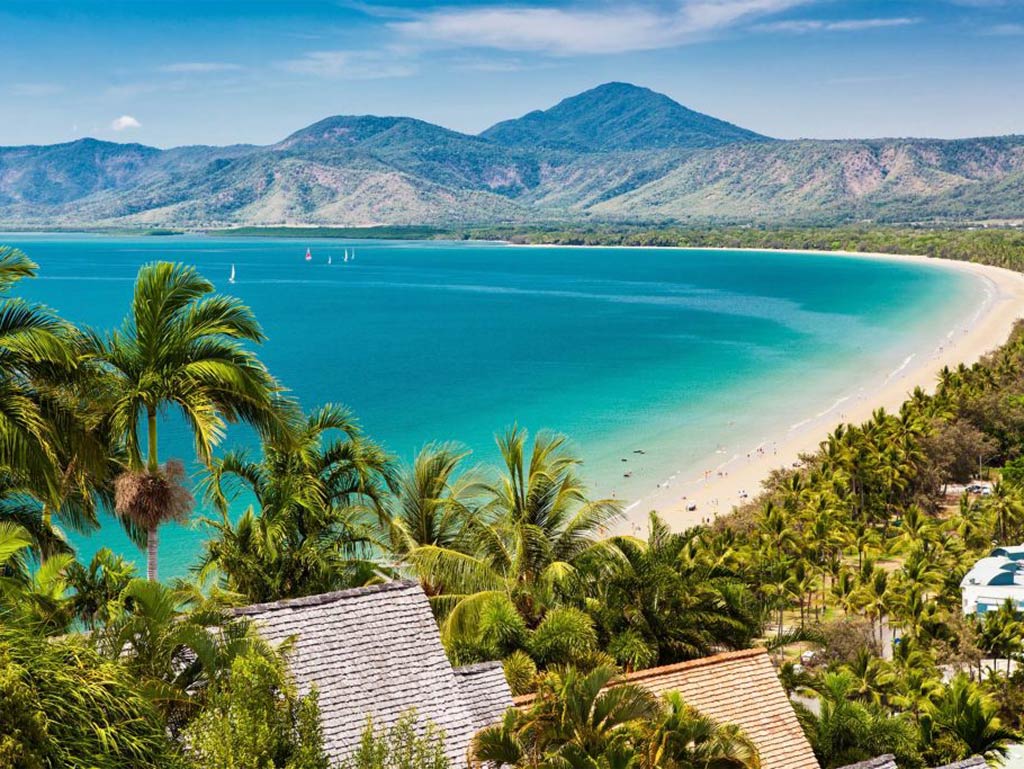 An aerial view of one of Cairns's beaches with mountains in the background and gorgeous turquoise water in front of it 