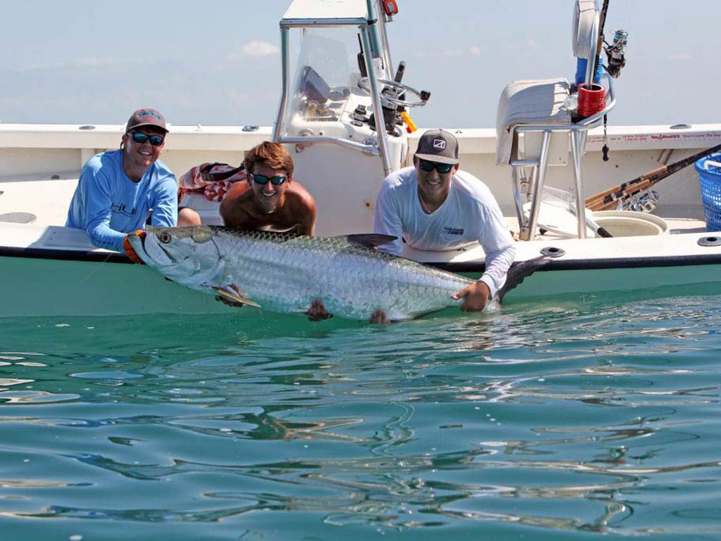Three anglers leaning from a boat, holding a Tarpon they caught just above the water