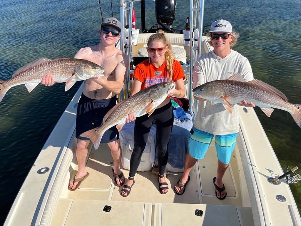 Three young anglers standing on a center console fishing boat, each holding a Redfish they caught.