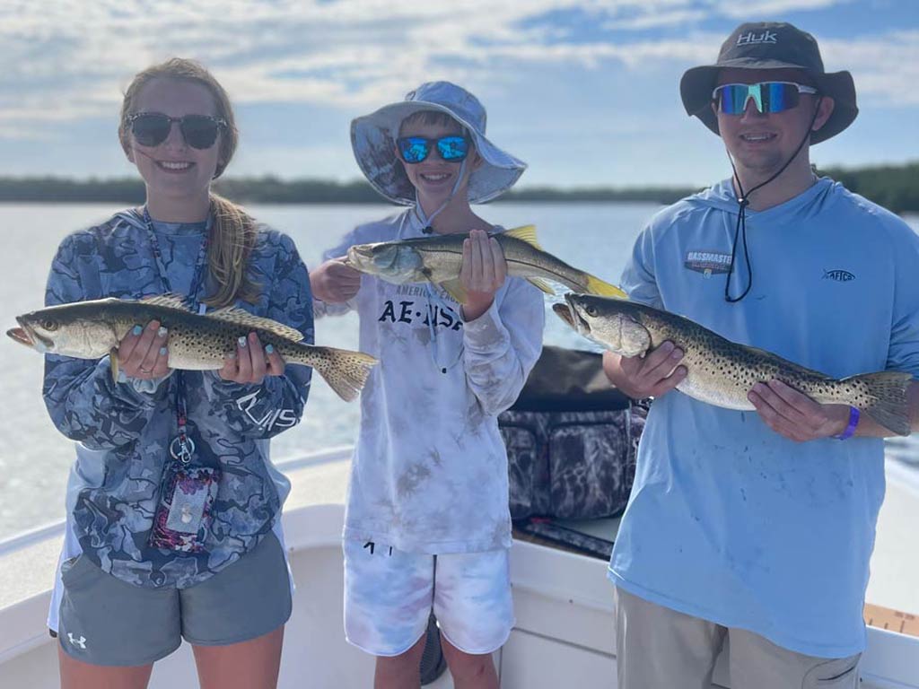 Three kids standing on a fishing boat, two of them holding a Spotted Seatrout, the one in the middle holding a a Snook.