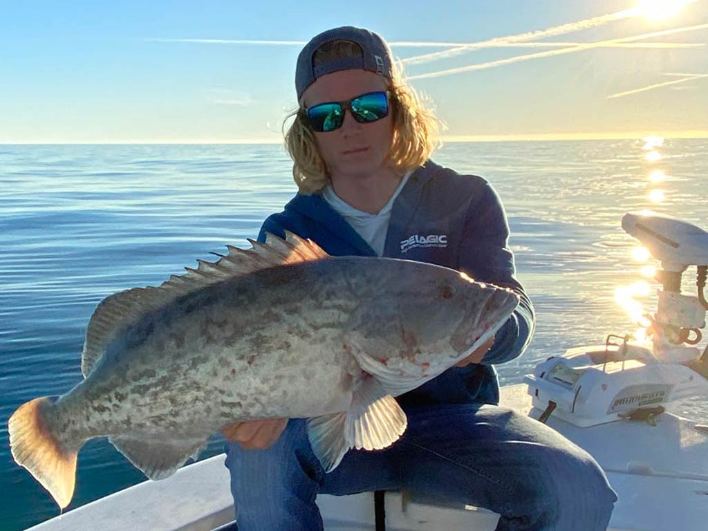 A young charter guide sitting on his fishing boat in a reversed cap and sunglasses, holding a Black Grouper he caught, with calm waters, blue skies, and the sun behind him.