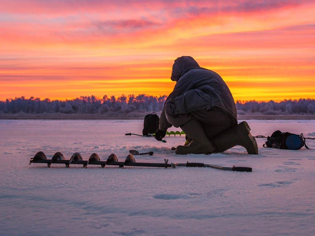 A photo featuring a dedicated ice fisherman drilling a hole on the frozen lake during the sunset