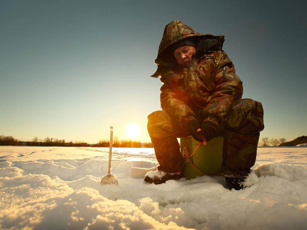 A photo featuring an ice fishing angler while sitting on a stool and carefully setting up his ice fishing post during the sunrise