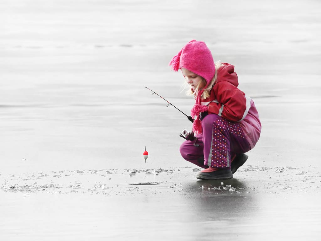 A photo featuring a little girl wearing a ping winter suit and a pink cap while squatting on a frozen lake next to a hole with an ice fishing rod