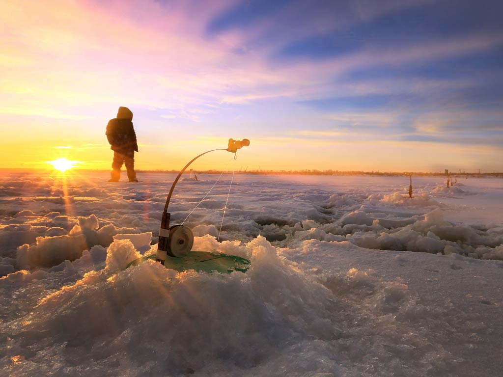 A photo featuring an ice fishing angler in the distance and an ice fishing gear and setup in front of him during the sunrise