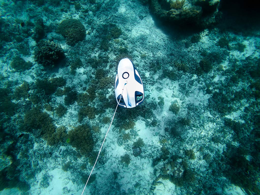 An underwater drone, designed for drone fishing, navigates over a coral reef. It's connected by a cable and surrounded by vibrant marine life.