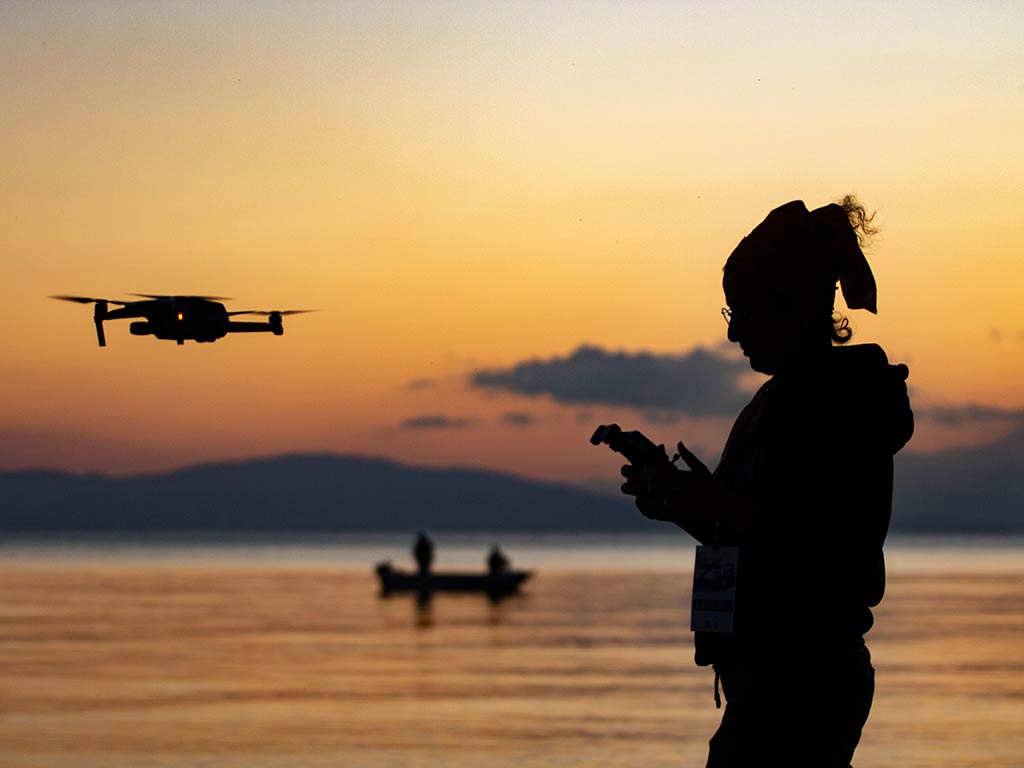 A silhouette of a woman who's engaged in drone fishing at sunset near a calm body of water, with a distant boat on the horizon.
