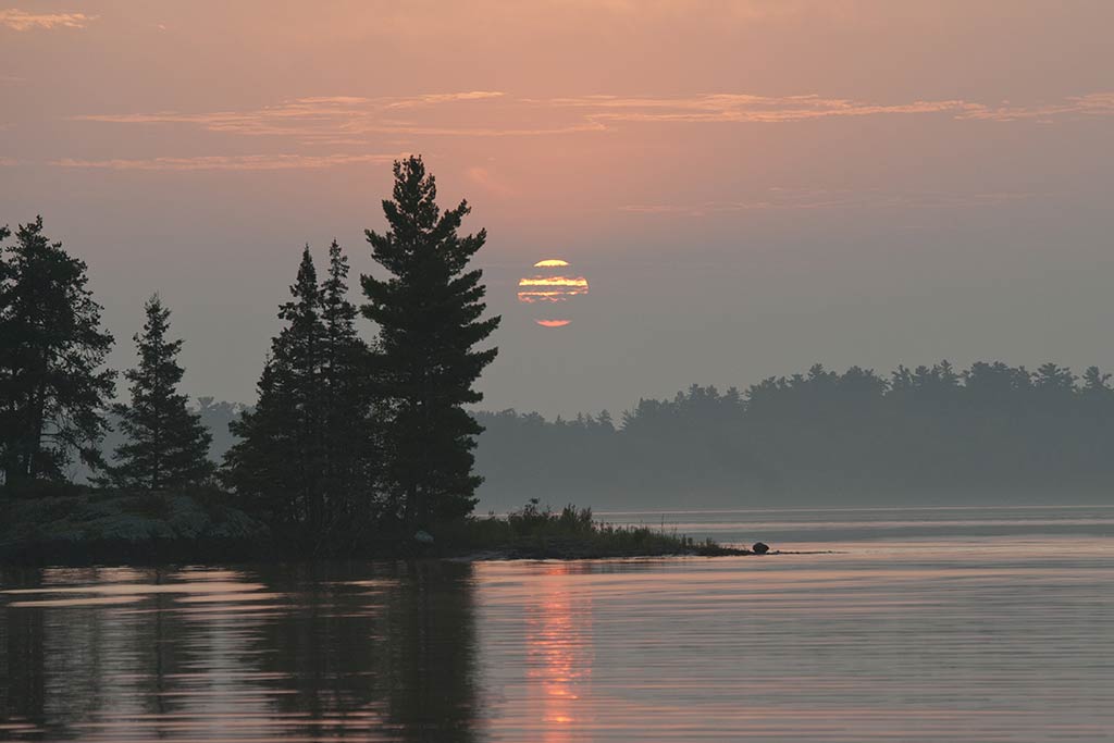 A view across the calm waters of Lake of the Woods in Canada looking towards the sun setting behind clouds and trees in the distance