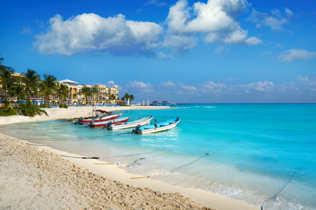 Four boats docked at a beach in Playa Del Carmen, looking out to the electric blue sea, with a resort behind them