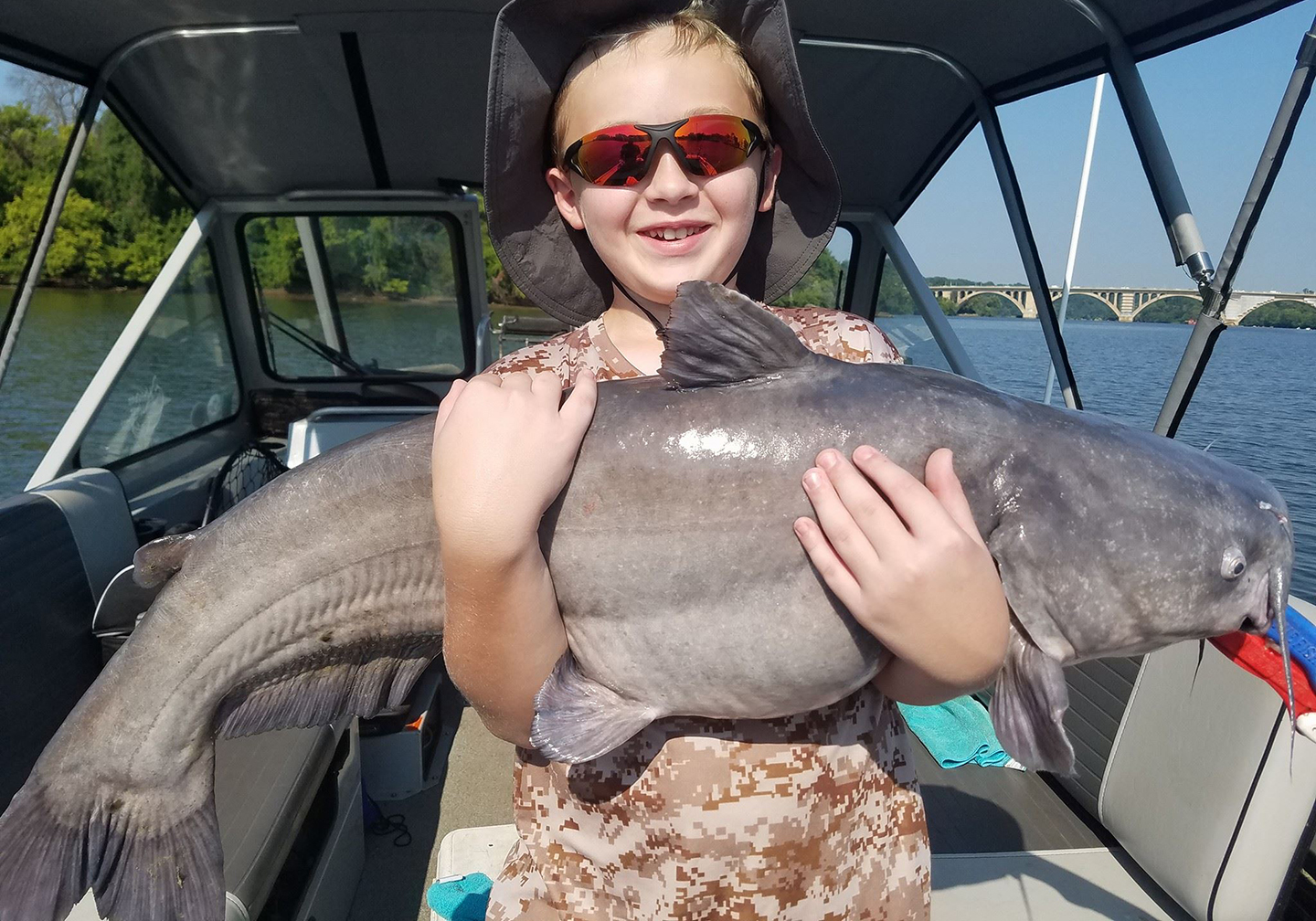 A happy child in sunglasses and a fishing hat, standing on a boat holding a big Blue Cat caught on a catfishing trip.