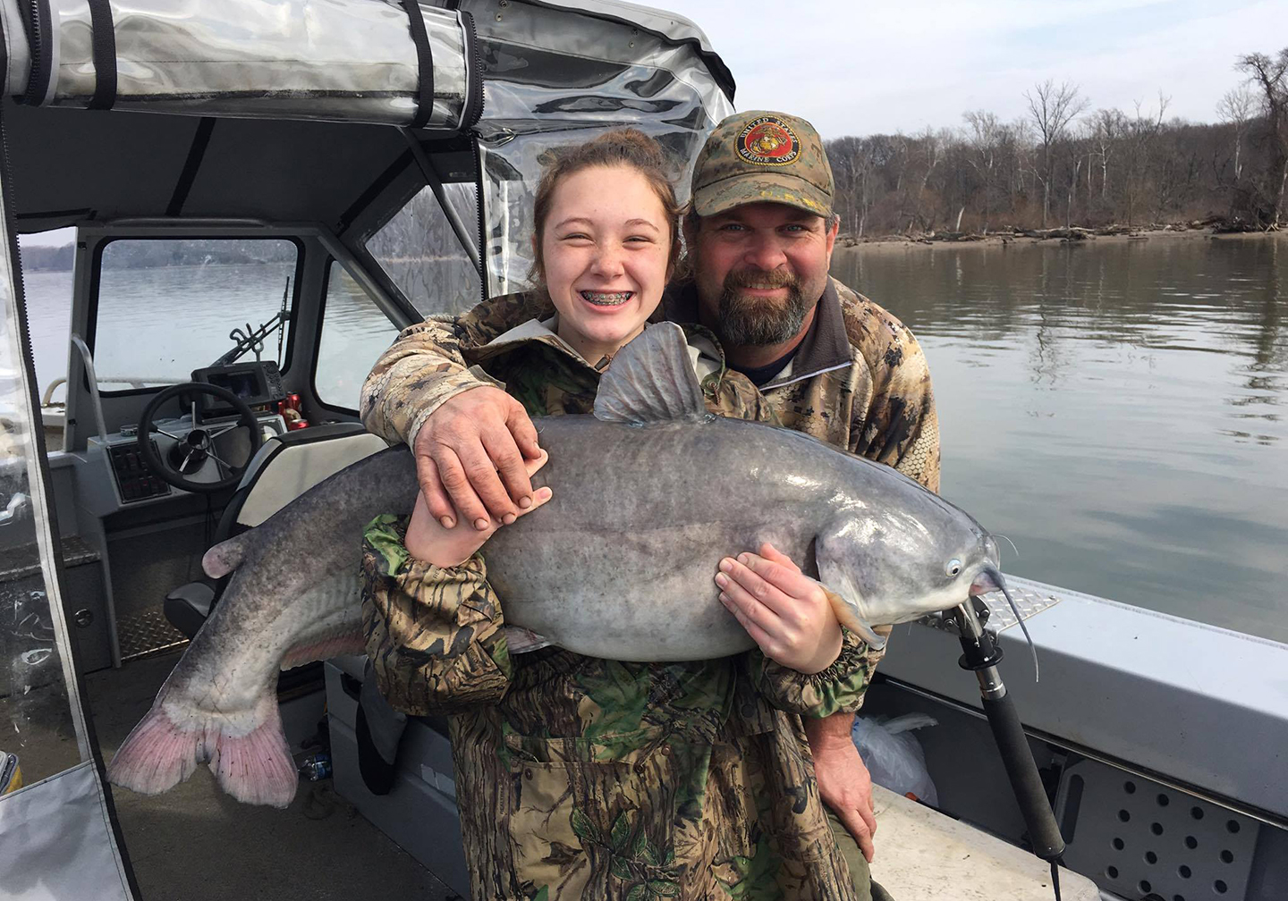 A man with his arm around a young girl who is holding a large Blue Catfish on a boat.