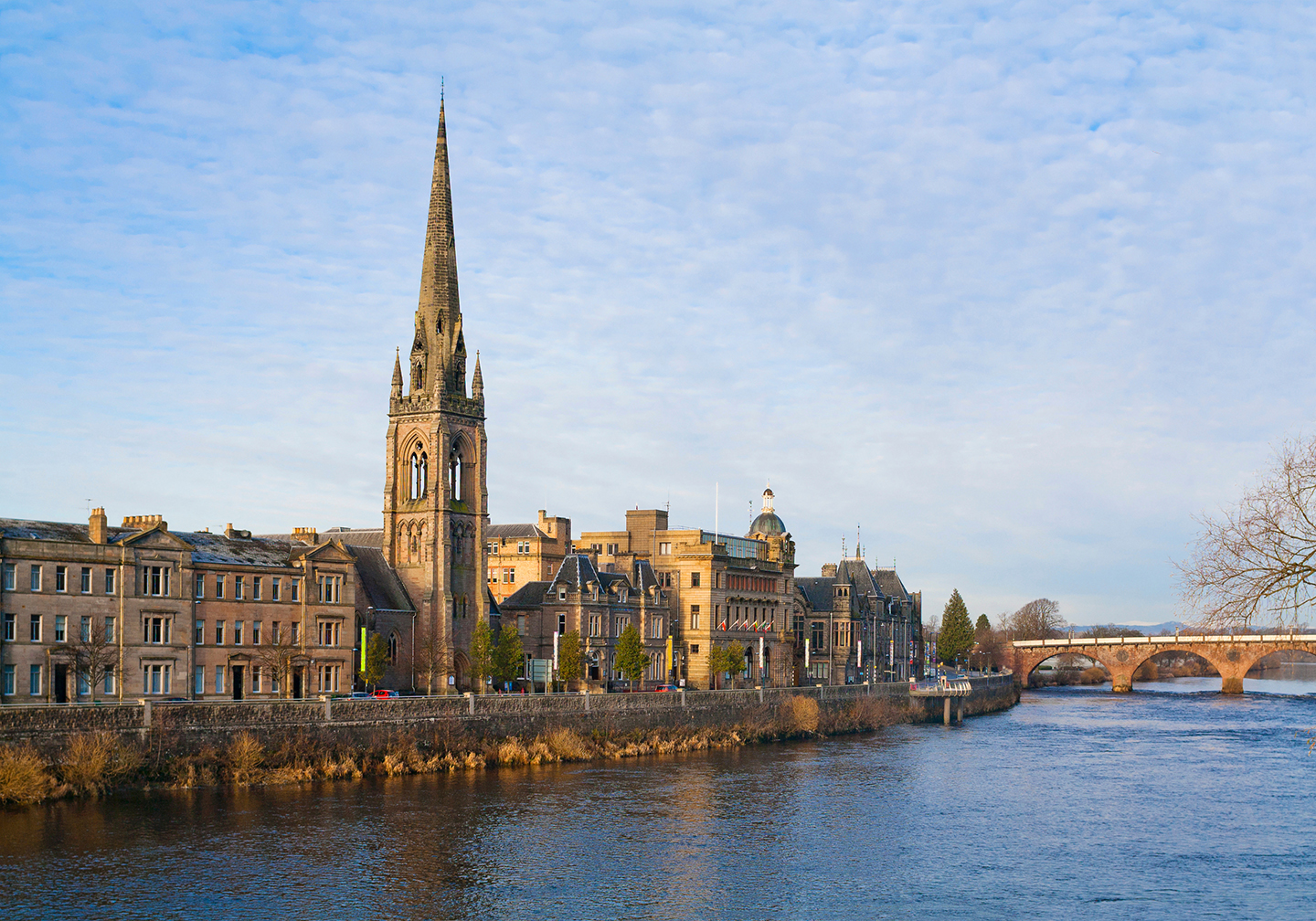 A view over the River Tay to the centre of Perth, Scotland, with an old stone church spire in the middle and a bridge on the right.