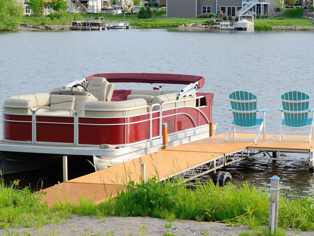 A photo of a red pontoon boat with white seats docked next to a wooden fishing pier and two blue chairs facing the water