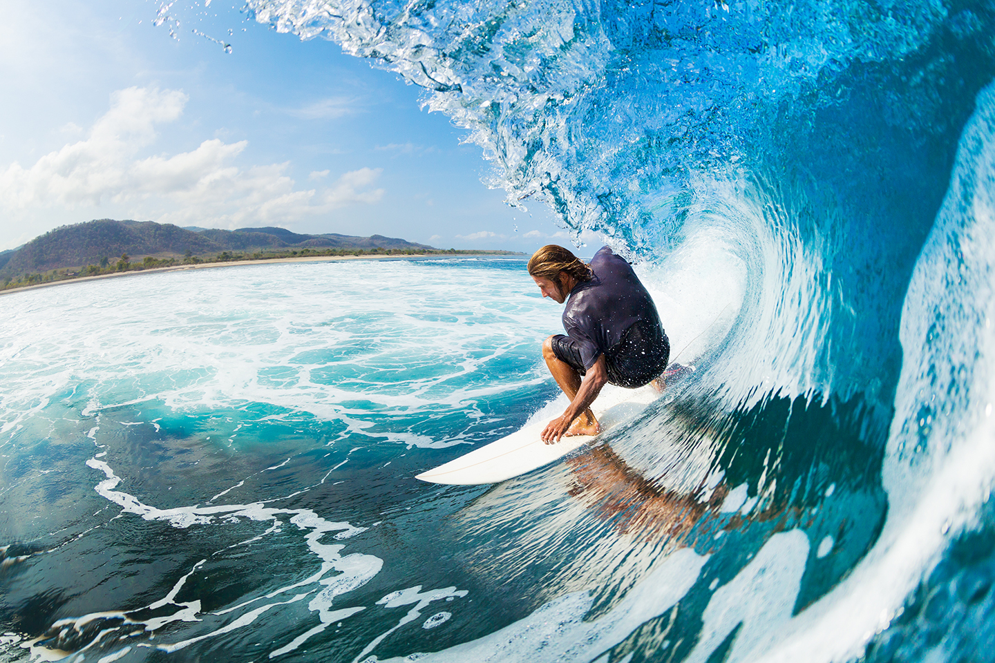 A man in a dark wetsuit surfing a large wave with the shore and mountains in the distance