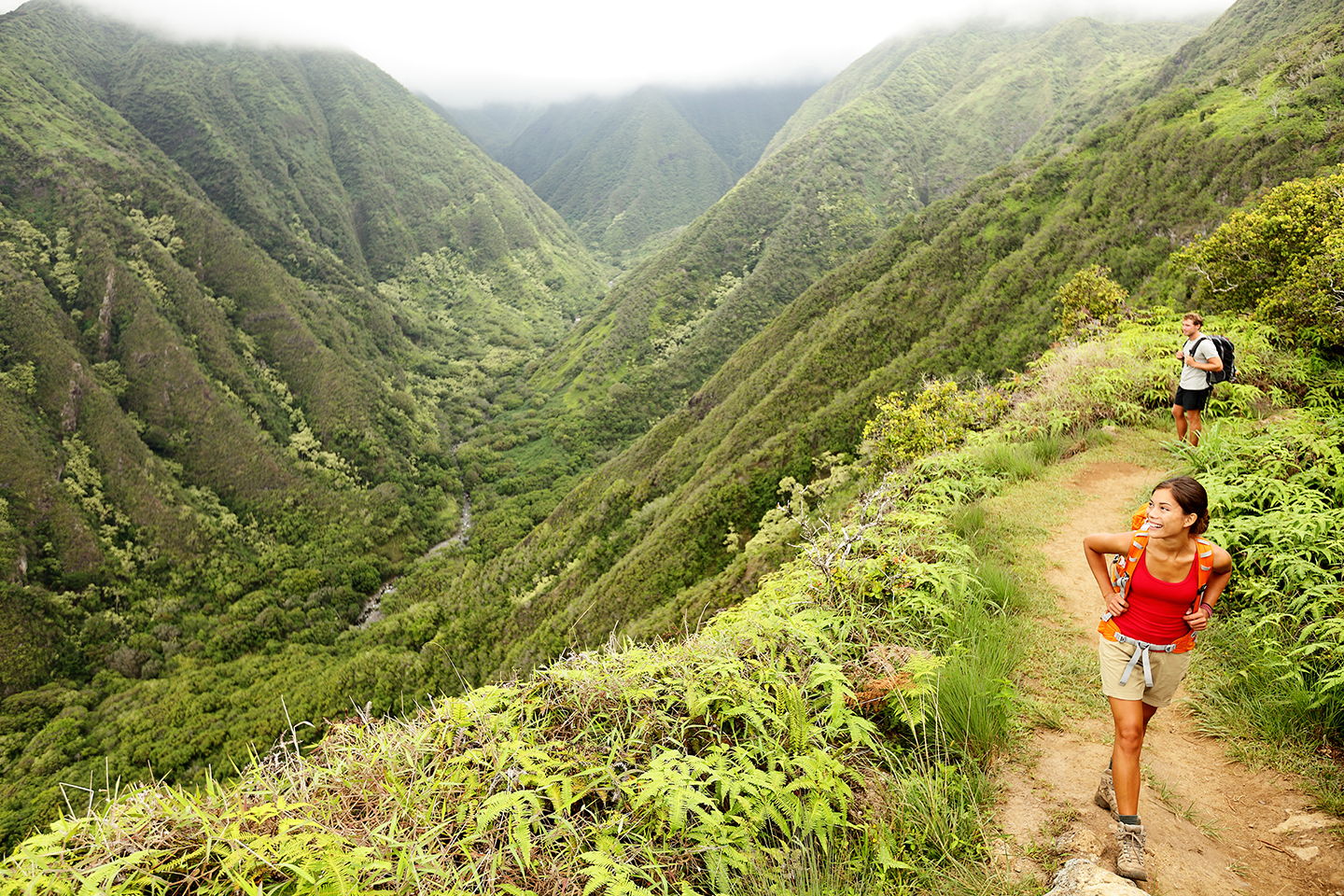 Two hikers walking along a mountain trail in Hawaii: a woman in a red tank top and beige shorts, and a man in a blue shirt and black shorts.