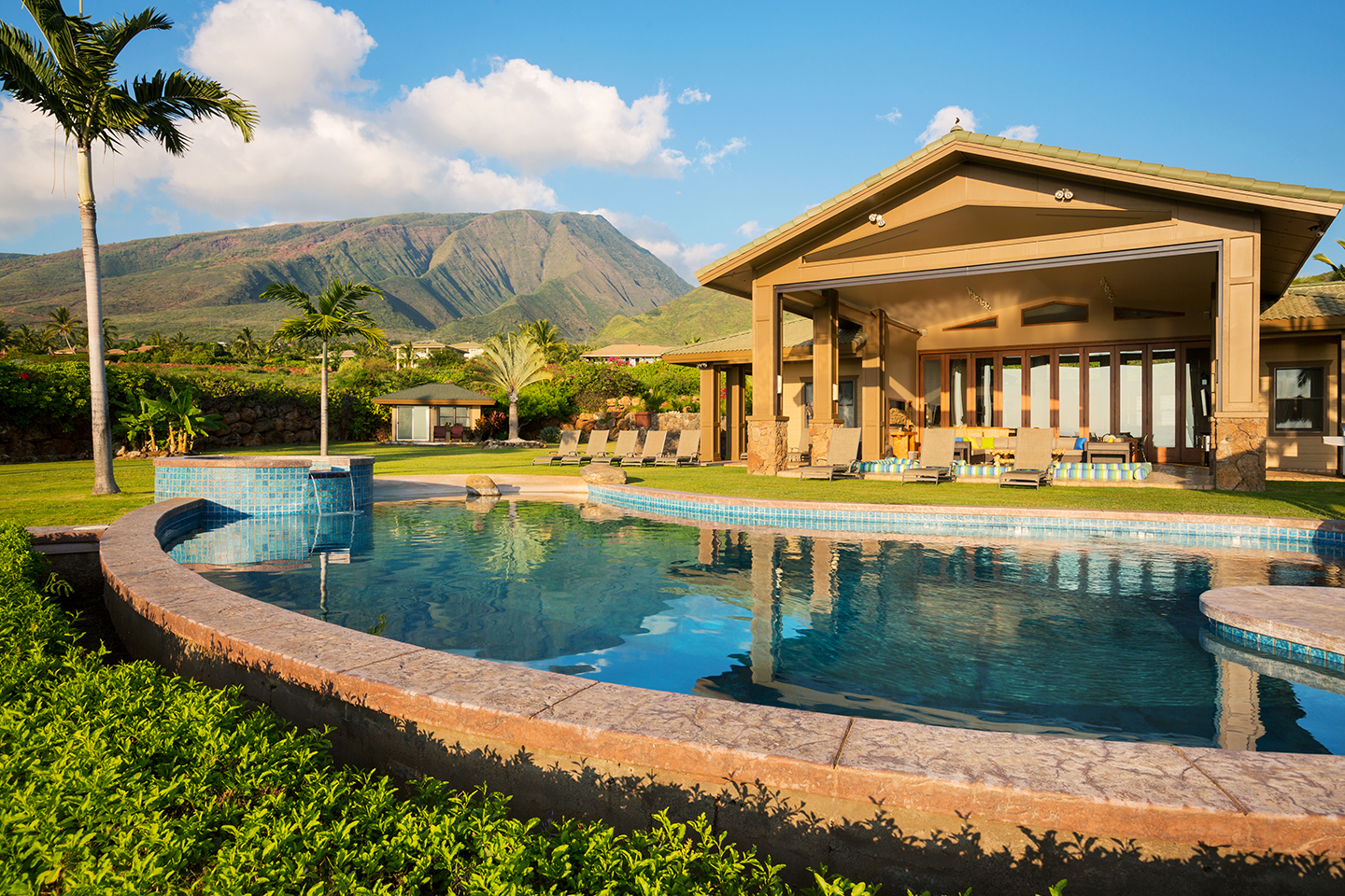 A luxurious tropical resort with a swimming pool, lawn, palm trees, and a mountain in the distance.