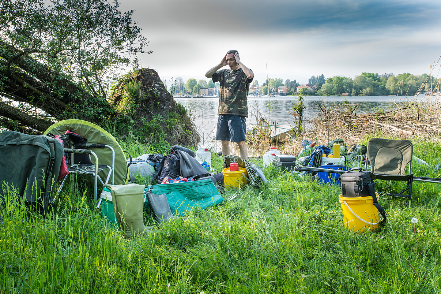 A carp fisherman looking panicked at the amount of fishing equipment surrounding him, which he has to pack and carry away from a lake after a day of fishing.