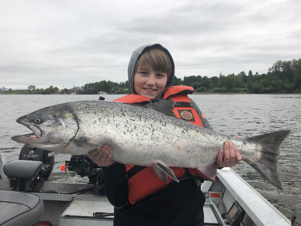 A child in an orange life jacket proudly showing a large Salmon, with cloudy sky and shimmering water as the perfect backdrop to this memorable catch.