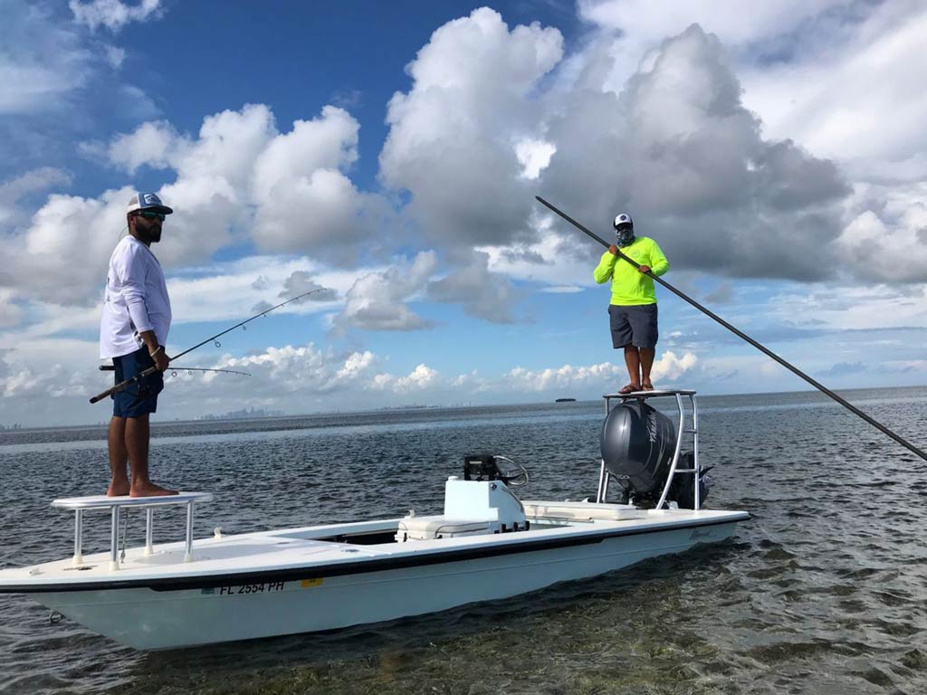 Two fly anglers standing on a boat in Miami flats, with one holding a long pole and another holding two fishing rods