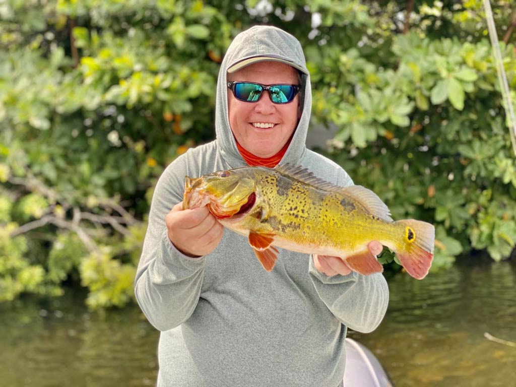 A middle-aged smiling angler in sunglasses, a cap and with a hood on, holding a Peacock Bass he caught, with waters and greenery behind him