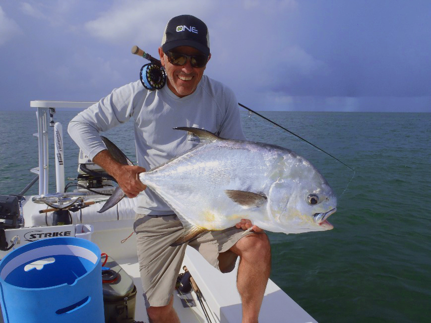 An angler in sunglasses and a hat kneeling on a boat and holding a Permit over his knee, with his fly fishing rod over his shoulder