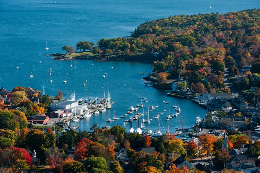 An aerial view of Camden, Maine, with boats in the water and autumn trees to the bottom and right