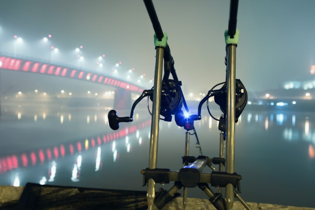 Two fishing rods set up by a river in a city at night. The lights of a nearby bridge are visible in the distance
