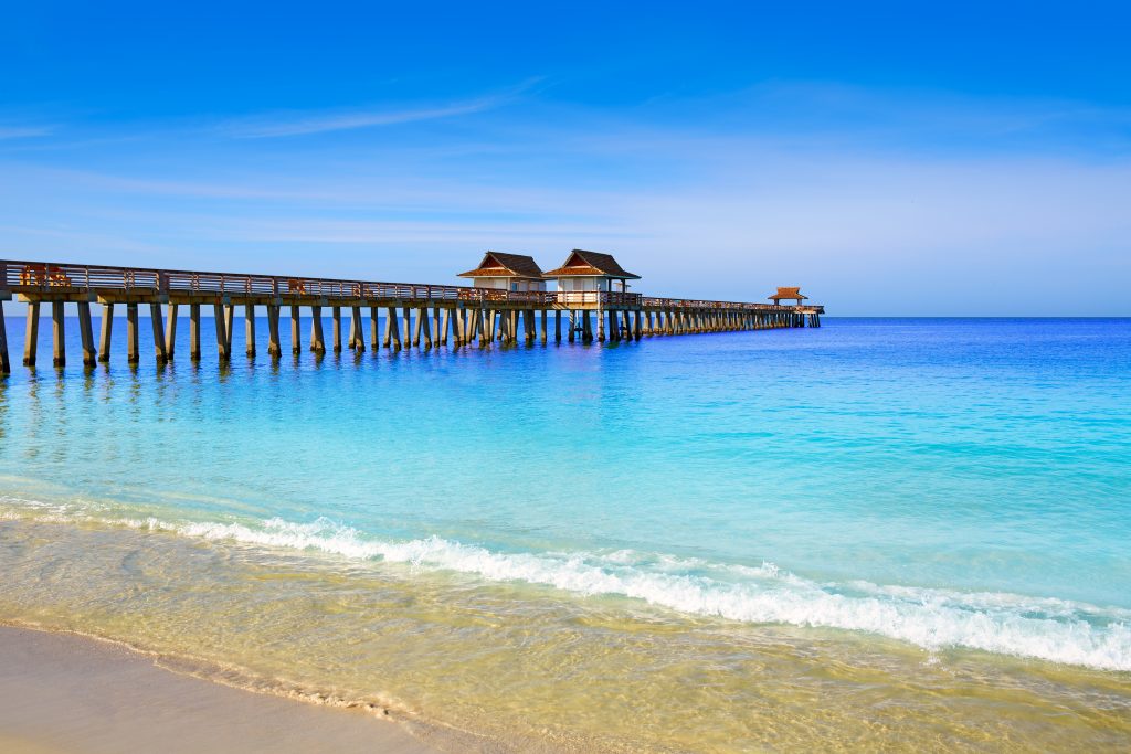 A view of Naples Pier from the beach, with bright blue water and yellow sand in the foreground