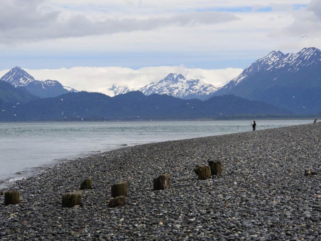 A photo of an angler fishing from shore on the Spit beach in Homer facing the magnificent snow-capped mountains in the distance