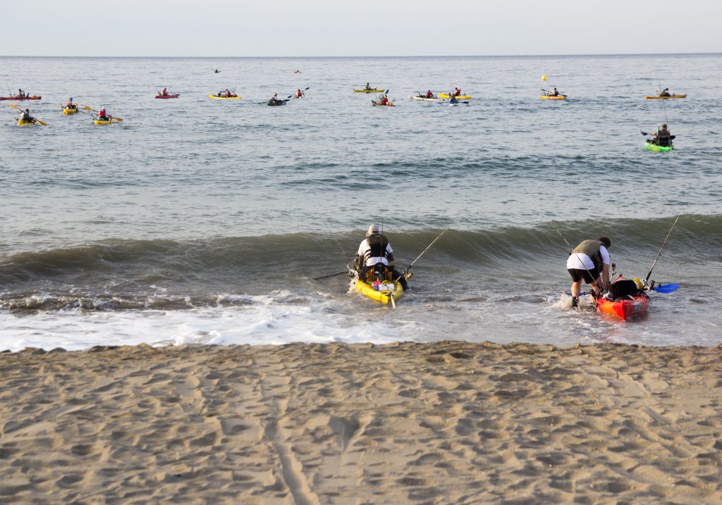 Two kayak anglers launching into the sea, with more fishing kayaks in the water ahead of them, as seen from the beach