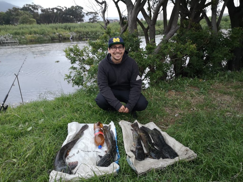 A man in a cap and glasses squatting behind his catch of Catfish on the bank of a river