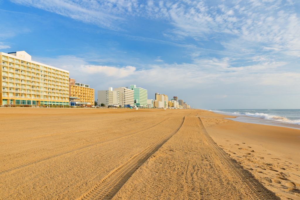 Virginia Beach in the early morning. A tyre trail stretches across the sand into the distance and hotels line the beachfront on the left.