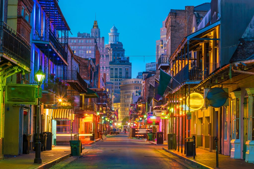 A narrow street in the French Quarter of New Orleans at dusk, with neon signs lighting the street.