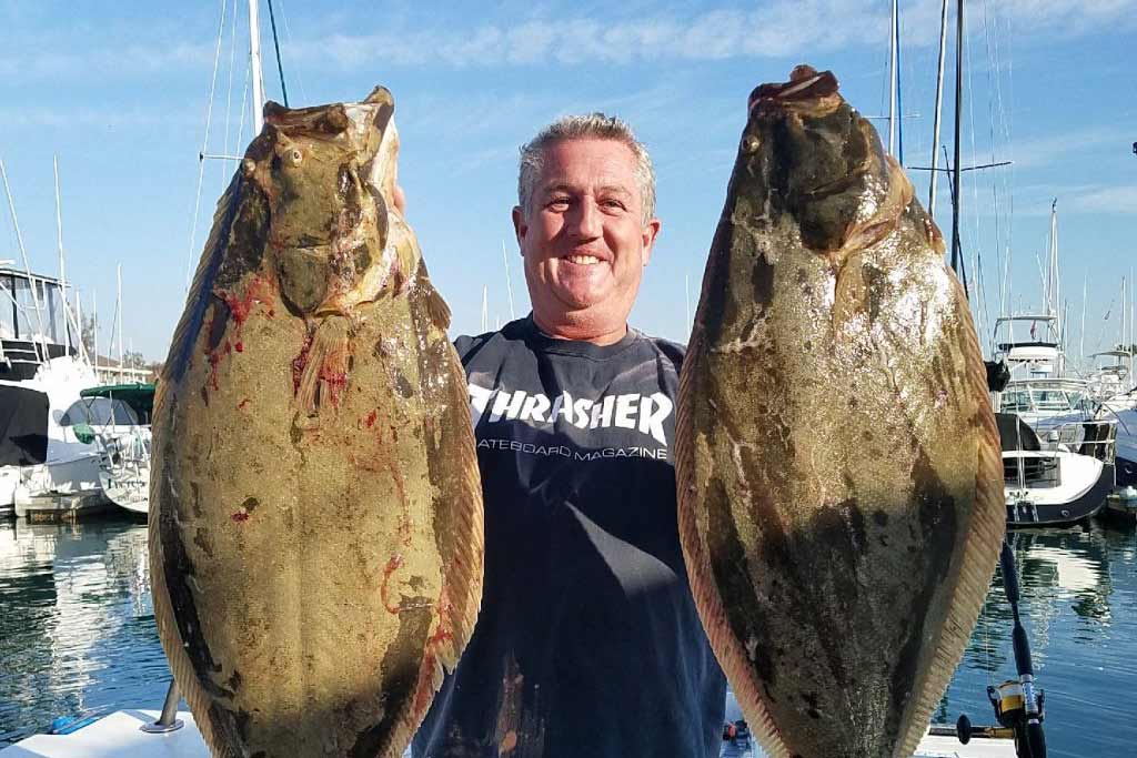 A middle-aged angler smiling widely while standing on a dock and holding two large Halibut he caught on his spring break fishing trip