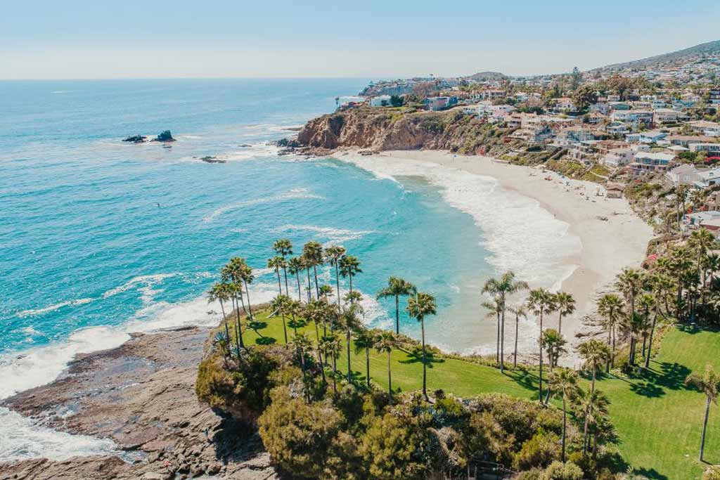 An aerial view of Laguna Beach, its clear, turquoise waters, pristine sand, and shoreline cliffs and buildings looking out to the sea