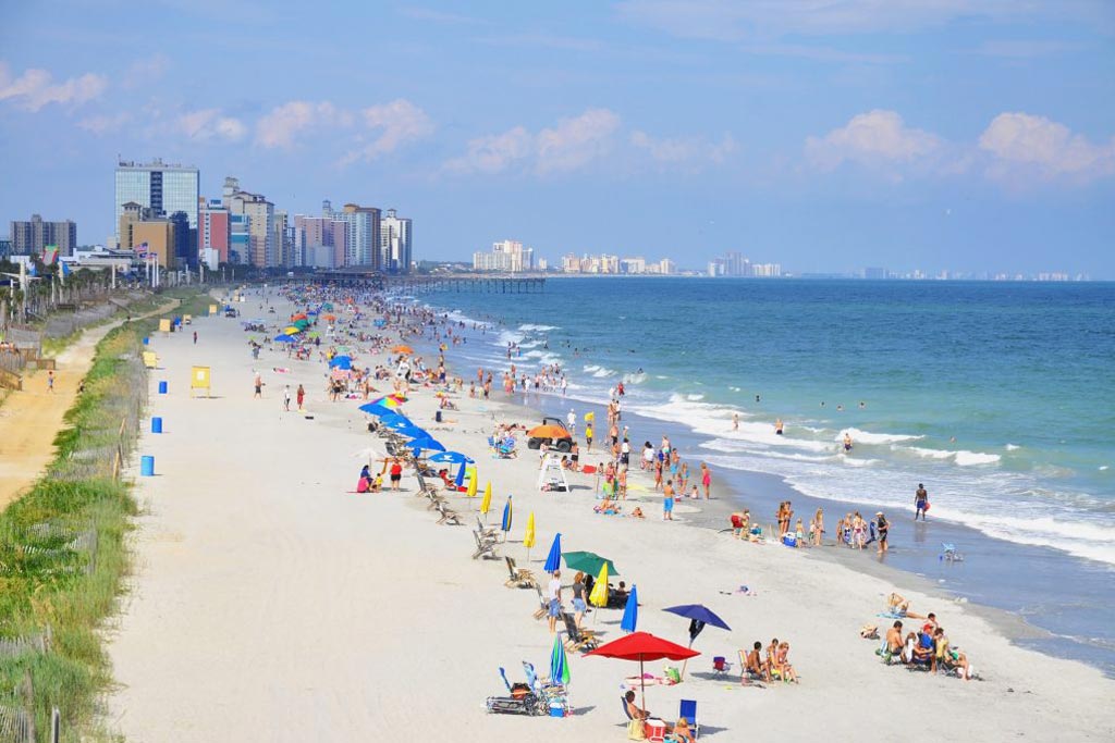 A photo of a beach on Myrtle Beach and the many people on it, in the distance there's a fishing pier and city buildings