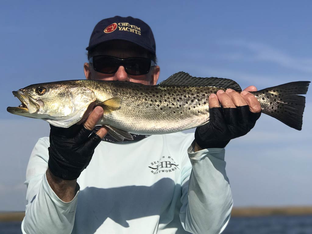 An angler holding a Spotted Seatrout with both hands in front of his face so that we can only see the fish and his sunglasses and a cap