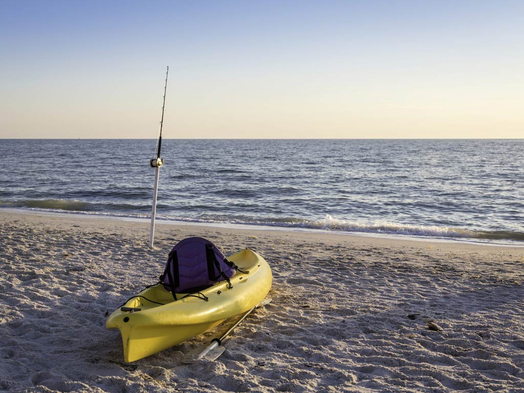 A yellow fishing kayak on a beach, with a surf fishing rod near the water behind it