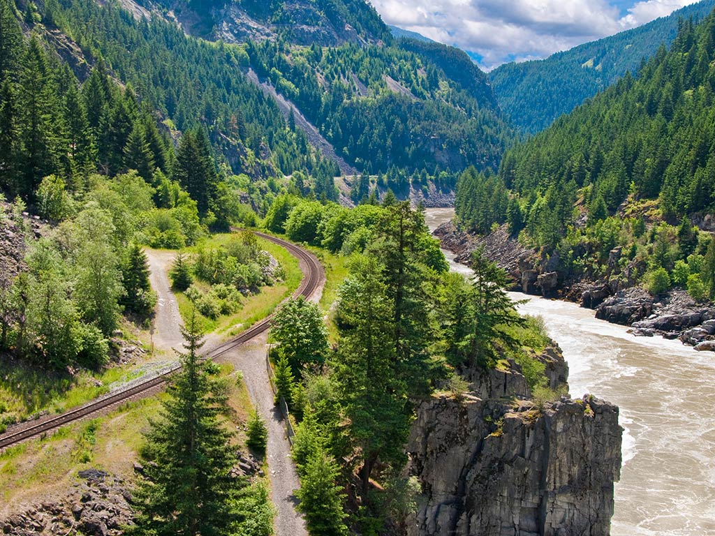 A view of the middle protion of the Fraser River as it winds its way around a gorge on a sunny day, with w aroad visible high up on the left of the image