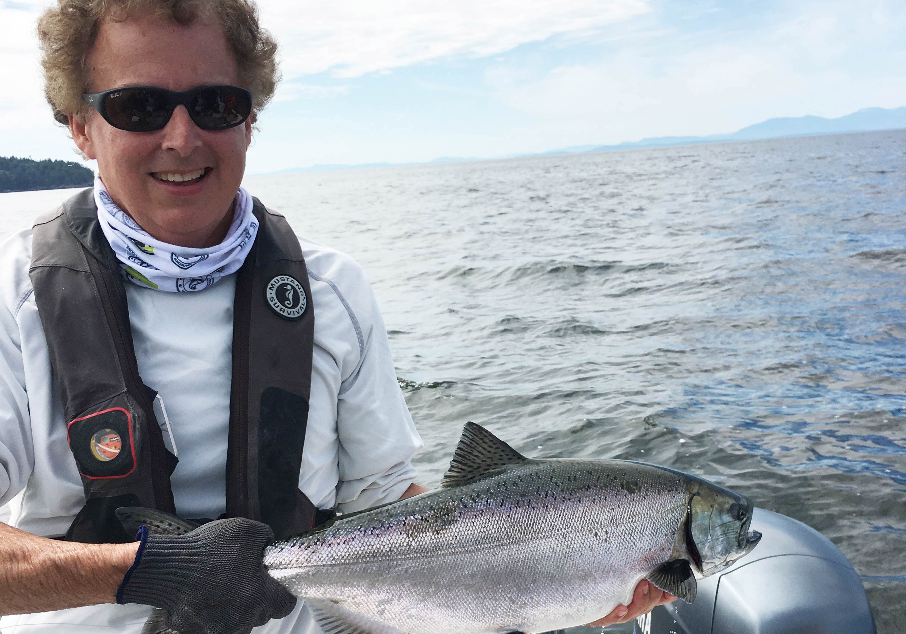 A man in sunglasses smiling and holding a Chinook Salmon with water in the background