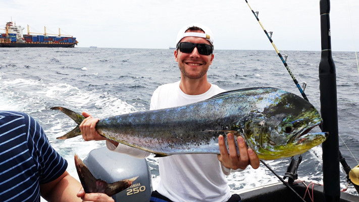 A happy angler in glasses, standing on a boat, holding a big Mahi Mahi with water in the background