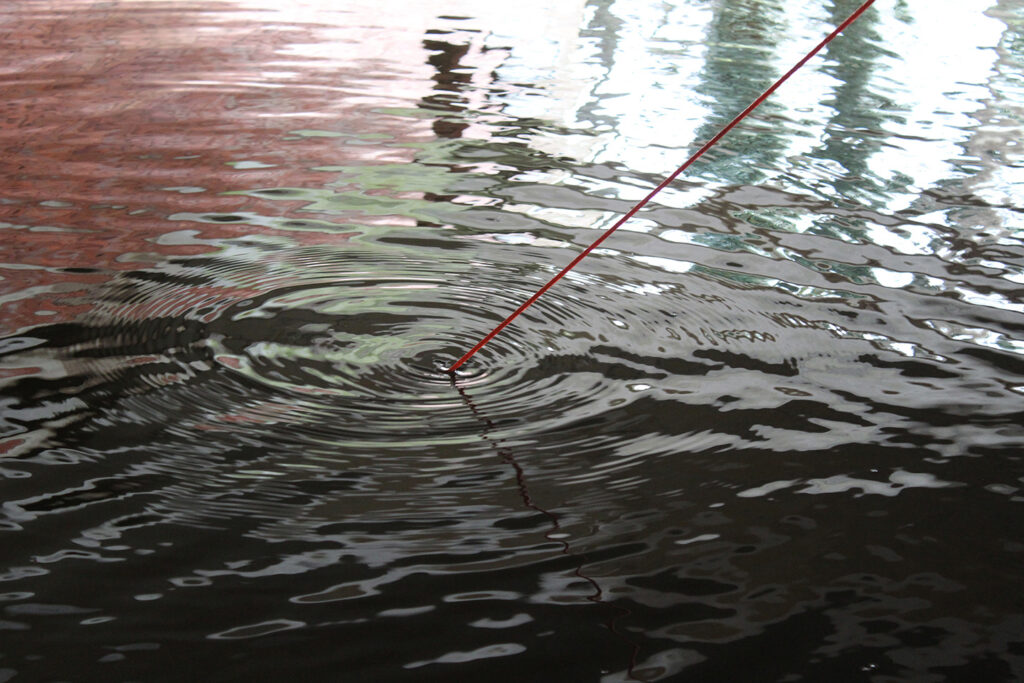 A thin red string for magnet fishing dips into rippling water, making circular waves. Reflections of a building or structure shimmer on the water&rsquo;s surface.