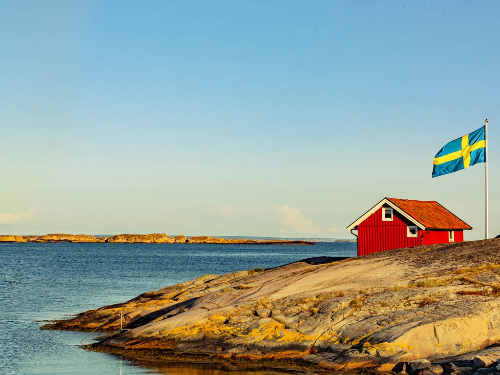 A red house next to a flag of Sweden both posing on the land on the verge with the lake during a bright and sunny day