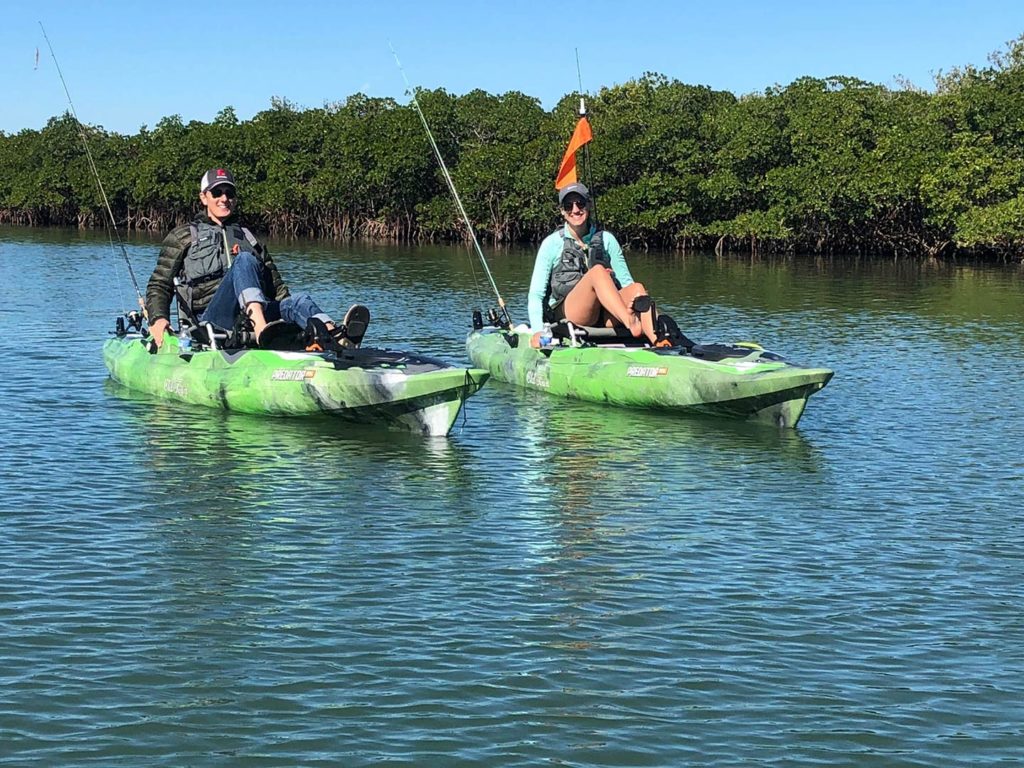 A male and female angler sit on their own green kayaks, pedaling their way to the inshore fishing grounds with greenery behind them