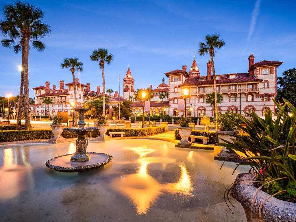 A view across the gardens towards Flagler College in St. Augustine, Florida at dusk, with palm trees and fountains lining the area before the impressive old building