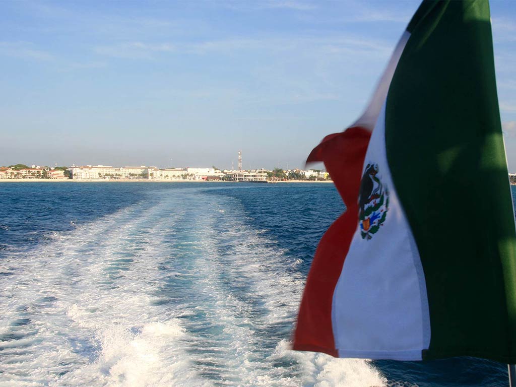 A Mexican flag flutters in the wind looking towards the coastline as a boat sails off into the sea