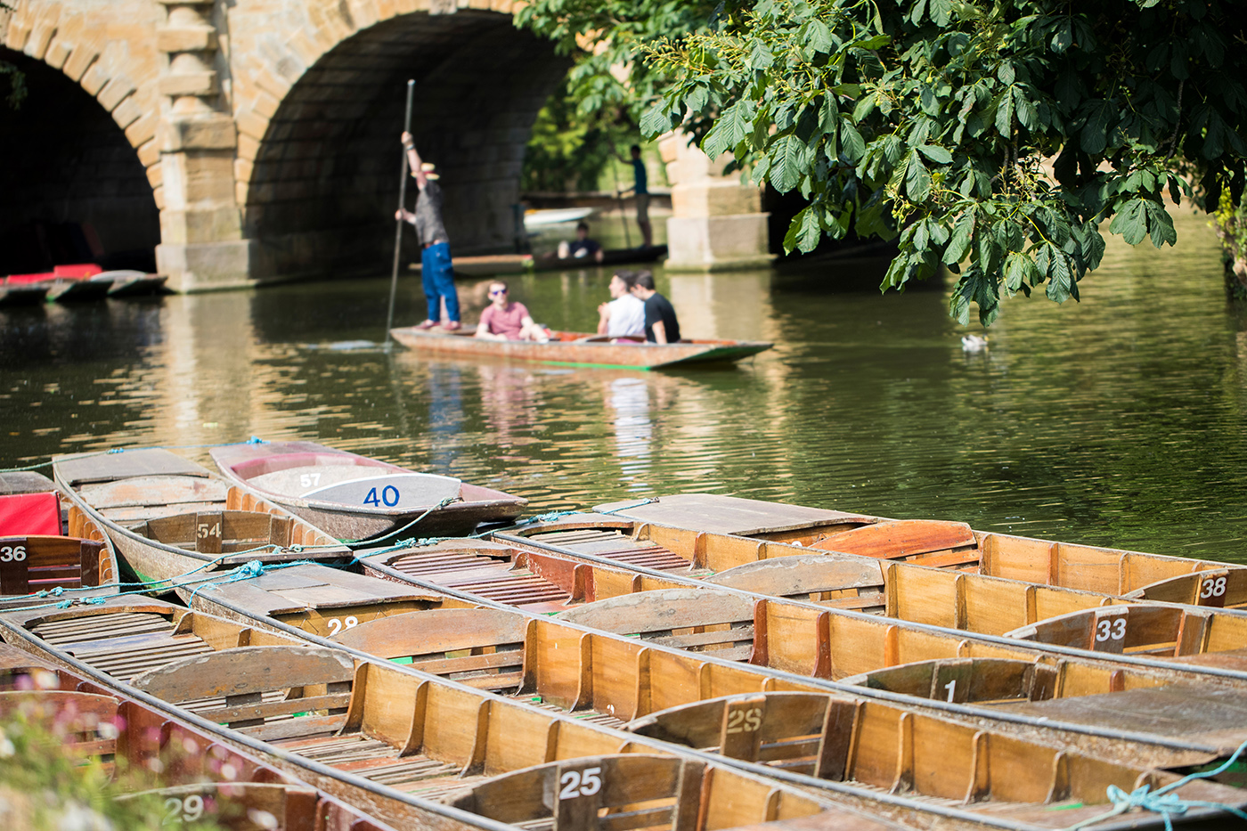 People punting on the river in Oxford, with boats in the foreground and Magdalen Bridge in the distance