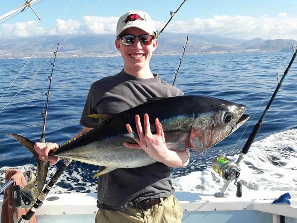 An angler stands on a charter boat offshore and holds a large Tuna with the ocean behind him