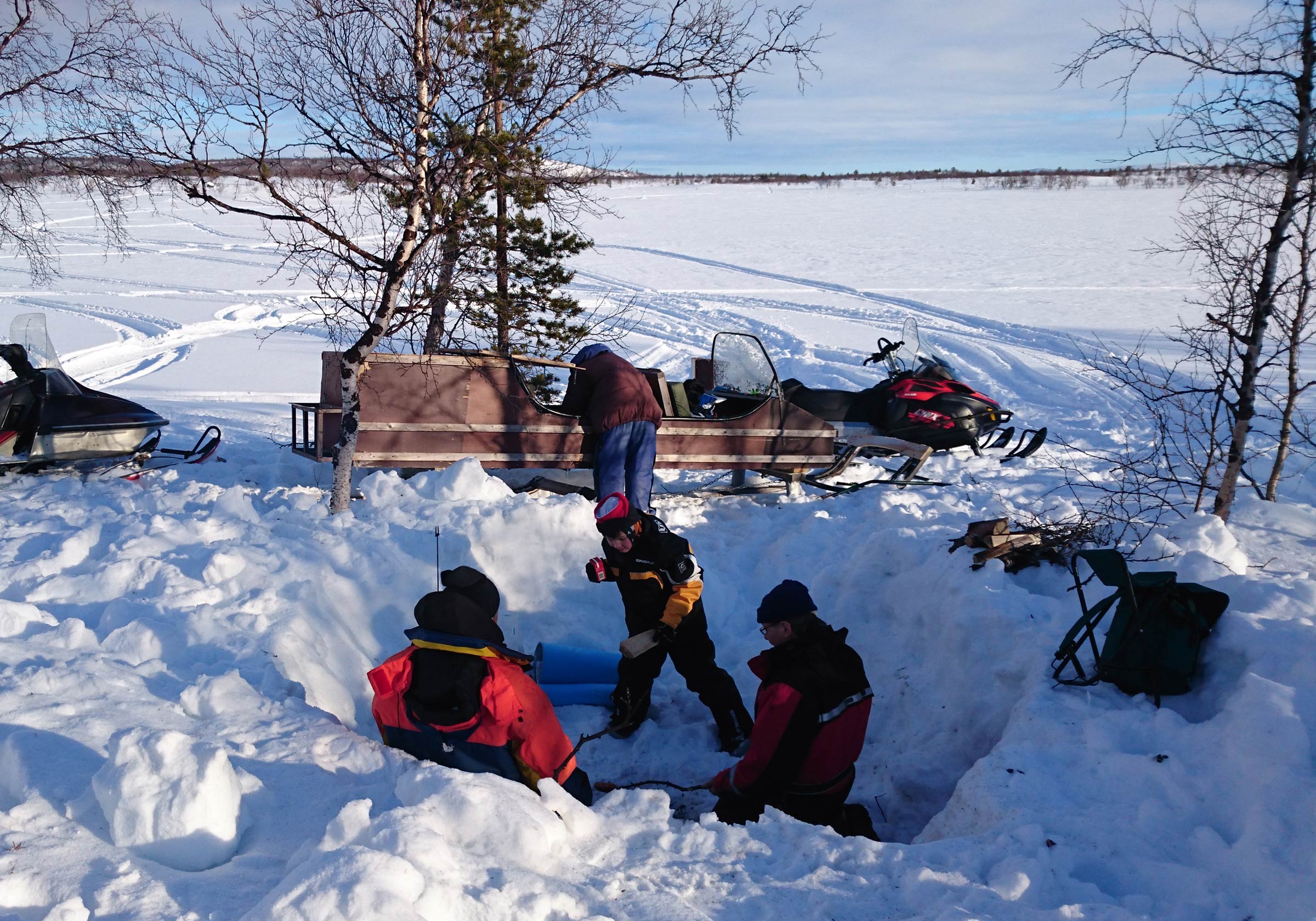 Four people in winter clothing are gathered in a snow pit near snowmobiles and trees on a snowy landscape under a partly cloudy sky.